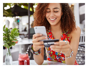 Beautiful dark skinned young female with cheerful expression, holds smart phone and credit card, banks online or makes shopping while sits against cafe interior. Payment and leisure concept.